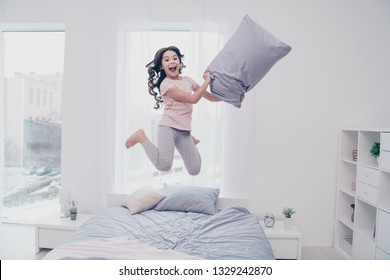 Full length body size view photo funky careless she her little girl jumping high bed  playful curly wavy pillow hands wear home t-shirt pants comfortable apartments flat bright light colored room