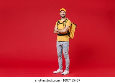 Full length body smiling delivery employee man in yellow cap t-shirt uniform thermal food bag backpack work courier service during quarantine covid-19 virus standing isolated on red background studio