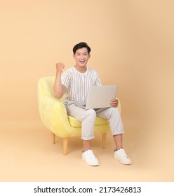 Full Length Body Stock Photo Of A Handsome Asian Young Man Sit On Yellow Sofa Chair, Isolated On Beige Background. Concept For App User, High Tech Gadgets Or Internet Promotion.