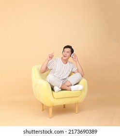 Full Length Body Stock Photo Of A Handsome Asian Young Man Sit On Yellow Sofa Chair, Isolated On Beige Background. Concept For App User, High Tech Gadgets Or Internet Promotion.