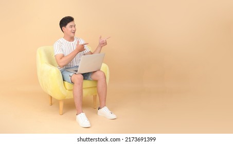 Full Length Body Stock Photo Of A Handsome Asian Young Man Sit On Yellow Sofa Chair, Isolated On Beige Background. Concept For App User, High Tech Gadgets Or Internet Promotion.