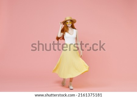 Full length body young nice smiling happy redhead woman 20s ginger long hair wear straw hat glasses summer clothes flow yellow maxi pleated skirt isolated on pastel pink background studio portrait