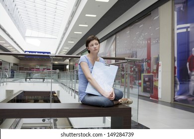 Full Length Of Bored Young Woman With Paper Bag Sitting In Shopping Centre