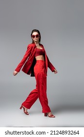 full length of brunette woman in red suit and trendy sunglasses walking on dark grey - Shutterstock ID 2124893264
