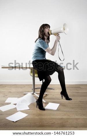 Full length of businesswoman screaming into megaphone while sitting on desk in office