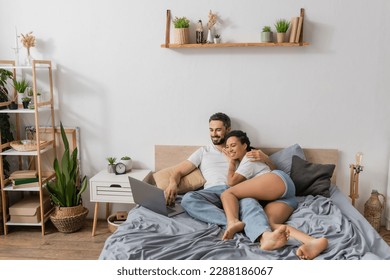 full length of carefree multiethnic couple watching film on laptop in modern bedroom at home