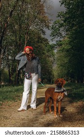 Full length of carefree woman in denim jacket and red beret standing with Rhodesian Ridgeback dog on pathway in park and enjoying sunny day in spring 