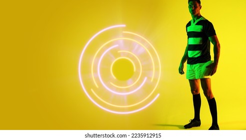 Full length of caucasian rugby player with ball standing by illuminated circular pattern, copy space. Yellow, composite, sport, competition, playing, match, shape and abstract concept. - Powered by Shutterstock - Get 15% off with code: PIKWIZARD15