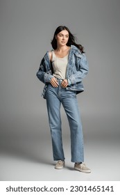 full length of charming young model with brunette hair posing while standing in stylish blue jeans and denim jacket and looking away on grey background