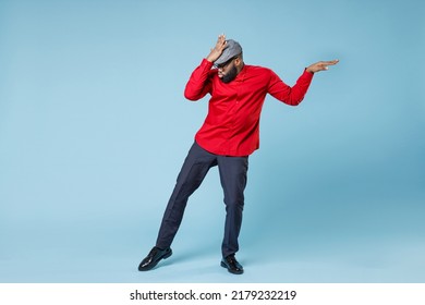 Full length of cheerful amazed young bearded african american man 20s wearing casual red shirt cap eyeglasses posing dancing put hand on head isolated on pastel blue wall background studio portrait