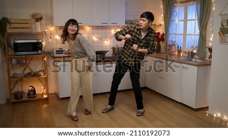 full length of cheerful asian couple doing funny dance moves to the music while dating celebrating valentine’s day in the kitchen at home in the evening