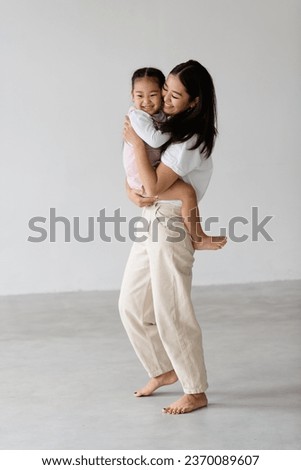 Full length of cheerful and barefoot young asian mom holding toddler daughter on grey background
