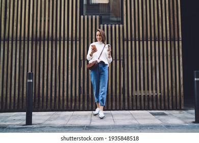 Full length of cheerful lady in trendy clothes chatting on smartphone and drinking takeaway coffee while spending time on city street - Powered by Shutterstock