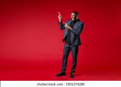 Full Length Of Cheerful Laughing Young African American Business Man 20s Wearing Classic Jacket Suit Standing Pointing Index Fingers Aside Isolated On Bright Red Color Wall Background Studio Portrait