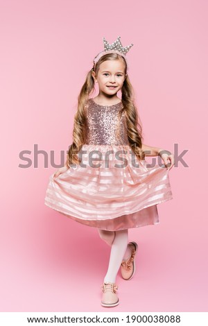 full length of cheerful little girl in dress and crown posing on pink