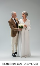 Full Length Of Cheerful Mature Bride In Wedding Dress Holding Bouquet And Standing With Middle Aged Groom In Suit On Grey