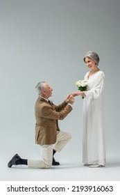 Full Length Of Cheerful Mature Man Standing On Knee And Wearing Engagement Ring On Finger Of Bride With Wedding Bouquet On Grey