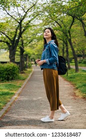 Full Length Of Cheerful Traveler Standing In The Path Looking Up To The Sky. Elegant Woman Enjoy The Beauty Of the Green Park In Summer. Asian Girl Tourist Travel In Osaka Lifestyle Concept.