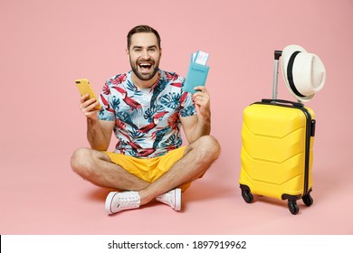 Full Length Cheerful Traveler Tourist Man Sit On Floor Using Mobile Phone Booking Hotel Taxi Hold Passport Tickets Isolated On Pink Background. Passenger Travel On Weekend. Air Flight Journey Concept