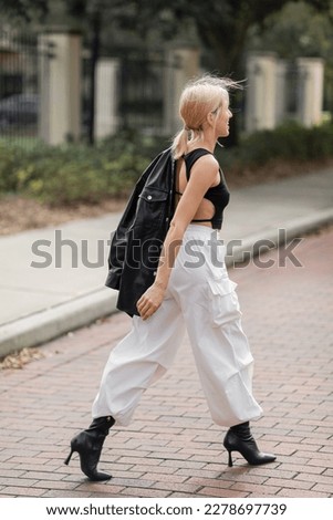 full length of cheerful woman in white cargo pants and boots walking with black leather jacket on street in Miami