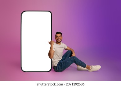 Full length of cheerful young Arab man sitting near big cellphone with mockup for mobile app on screen in neon light. Smartphone display template for new website or online ad