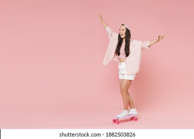 Full length of cheerful young asian woman in casual clothes, cap isolated on pastel pink background studio portrait. People lifestyle concept. Mock up copy space. Riding on skateboard walking have fun