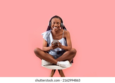 Full length of cheerful young black woman with headphones and cellphone listening to music or audio book, sitting on chair, using new mobile app on pink studio background - Powered by Shutterstock