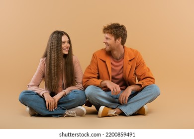 Full Length Of Cheerful Young Couple In Jeans And Jackets Sitting And Looking At Each Other On Beige