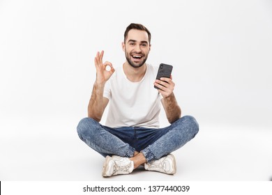 Full length of a cheerful young man sitting with legs crossed isolated over white background, using mobile phone, ok