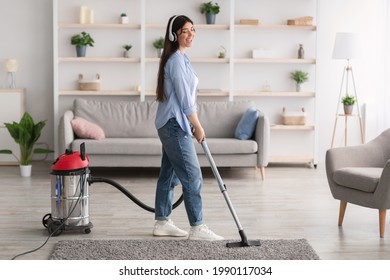 Full Length Of Cheerful Young Woman Listening To Music In Wireless Headphones While Vacuuming Her Apartment, Using Hoover, Indoors. Happy Millennial Lady Singing, Enjoying Domestic Chores