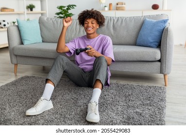 Full Length Of Cheery Black Adolescent Enjoying Videogame On Playstation, Having Fun At Home. Positive Afro Teenager With Joystick Sitting On Floor, Playing Computer Arcade. Lockdown Hobbies Concept