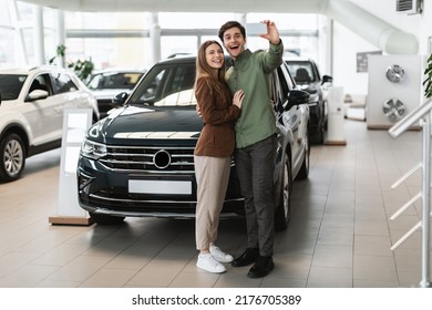 Full Length Of Cheery Young Couple Taking Selfie On Mobile Phone Near Their New Car At Auto Dealership, Copy Space. Joyful Millennial Spouses Photographing Themselves At Showroom, Buying Automobile