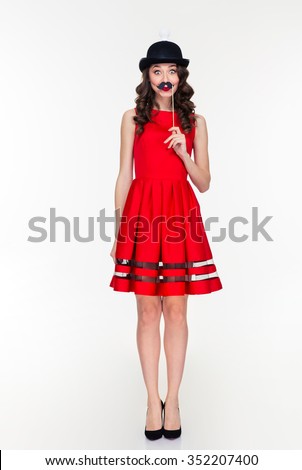 Full length of comical amusing young woman in red dress and ridiculous black hat with light bulb holding fake moustache on stick