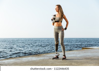 Full length of confident disabled athlete woman with prosthetic leg standing at the beach and drinking water from a bottle - Powered by Shutterstock