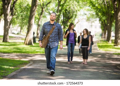 Full Length Of Confident Male Grad Student Walking On Campus Road With Friends In Background