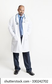 Full Length Confident Mature Indian Male Medical Doctor In Uniform Standing On Plain Background With Shadow.