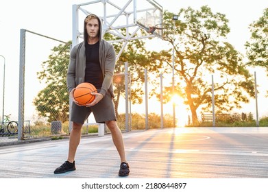 Full Length Confident Sunlit Young Sporty Sportsman Man In Grey Sports Clothes Hood Look Camera Training Hold In Hand Ball Play At Basketball Game Playground Court. Outdoor Courtyard Sport Concept.