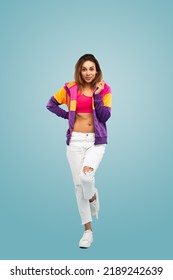 Full Length Of Confident Young Female Millennial With Long Brown Hair In Trendy Crop Top And Jacket, Standing Against Blue Background With Hand On Waist And Looking At Camera