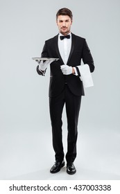 Full length of confident young waiter in tuxedo standing and holding tray over white background