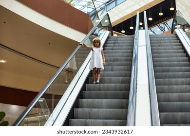 Full length of cute blonde little girl 5-6 year old in white dress going down escalator. Photo of adorable funny kid goes down, alone pensive looking. Happy joy childhood concept. Copy ad text space
