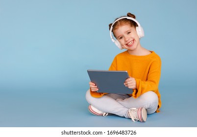 Full length delighted little girl in casual clothes and headphones smiling for camera and listening to music while sitting crossed legged and using tablet on blue background
 - Powered by Shutterstock