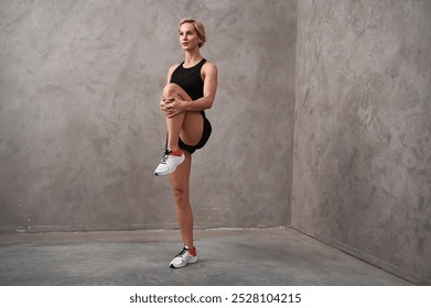 Full length of determined woman doing stretching exercise. Female trainer in a standing position pulls one leg up to the chest with the knee bent. - Powered by Shutterstock