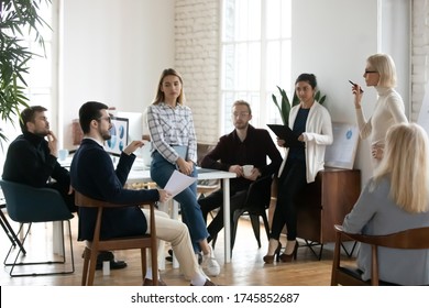 Full Length Different Ages Multiracial Businesspeople Listening To Confident Young Female Speaker Standing Near Flipchart. Smart Businesswoman Trainer Answering Questions Of Workshop Participants.