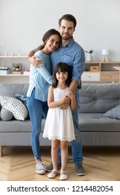 Full Length Diverse Multi-ethnic Family Married Couple Wife Husband Little Daughter Embracing Standing Together In Living Room Smiling Looking At Camera At New Modern Home Feels Happy And Satisfied