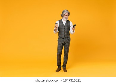 Full length of elderly gray-haired business man in checkered suit waistcoat shirt hold paper cup coffee or tea using mobile cell phone isolated on yellow background. Achievement career wealth concept