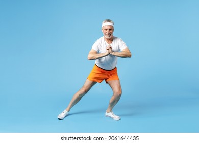Full length elderly gray-haired sportsman strong coach instructor man 60s in sportswear white t-shirt walk yoga mat do squats exercises warm-up isolated on blue background studio Fitness sport concept