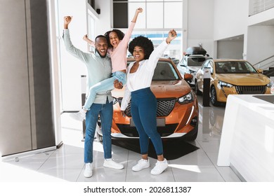 Full length of excited African American family lifting hands up, celebrating new car purchase at auto dealership. Black parents with daughter making YES gesture after buying vehicle at showroom