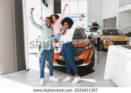 Full length of excited African American family lifting hands up, celebrating new car purchase at auto dealership. Black parents with daughter making YES gesture after buying vehicle at showroom