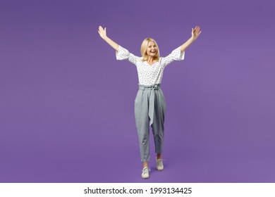 Full length of excited funny elderly gray-haired blonde woman lady 40s 50s in white dotted blouse standing rising spreading hands looking camera isolated on violet color background studio portrait