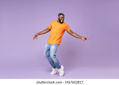 Full length of excited funny young african american man 20s in basic casual orange t-shirt dancing standing on toes pointing index fingers aside isolated on pastel violet background studio portrait